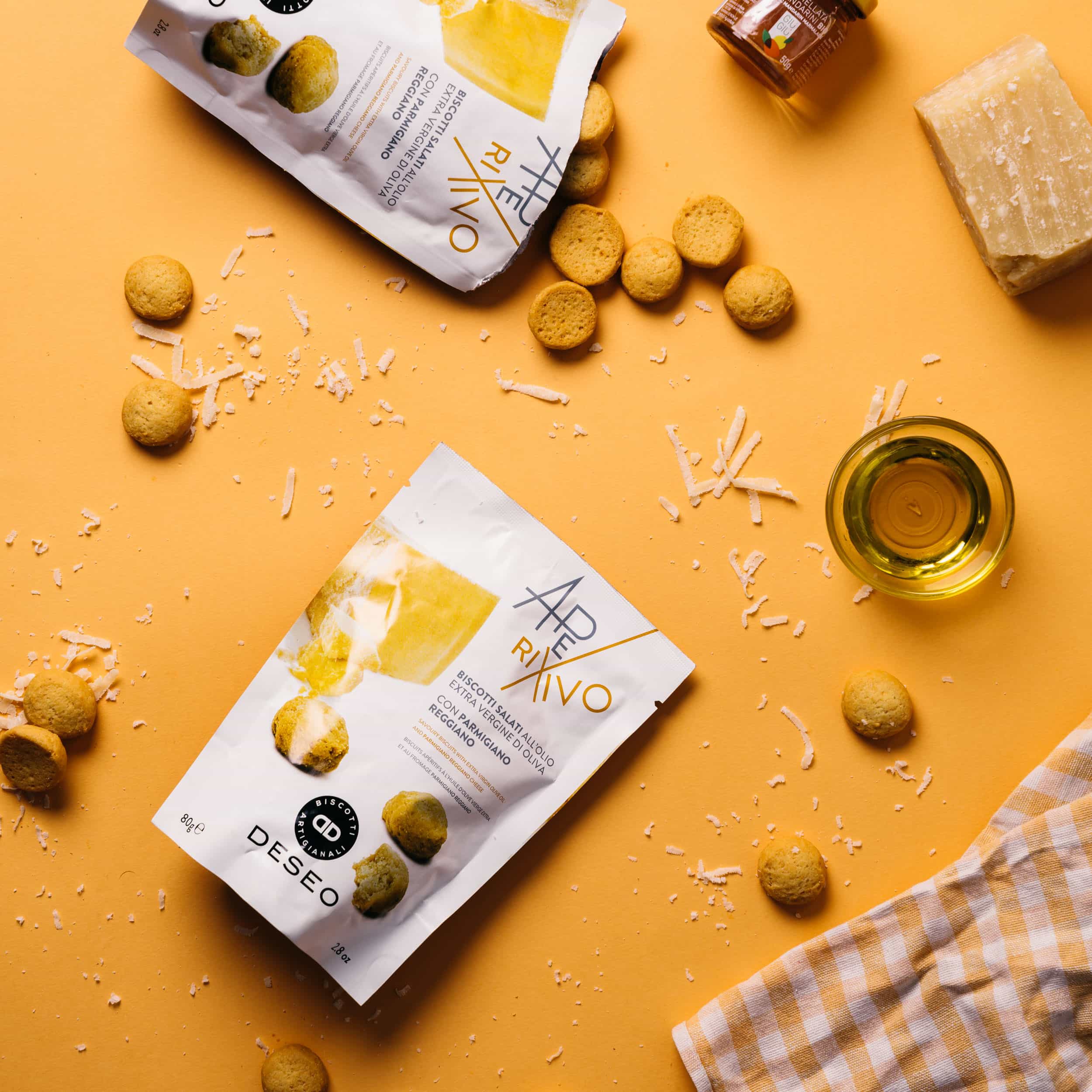 Two bags of Aperitivo snacks on a yellow surface with scattered snacks, grated cheese, and a glass of olive oil