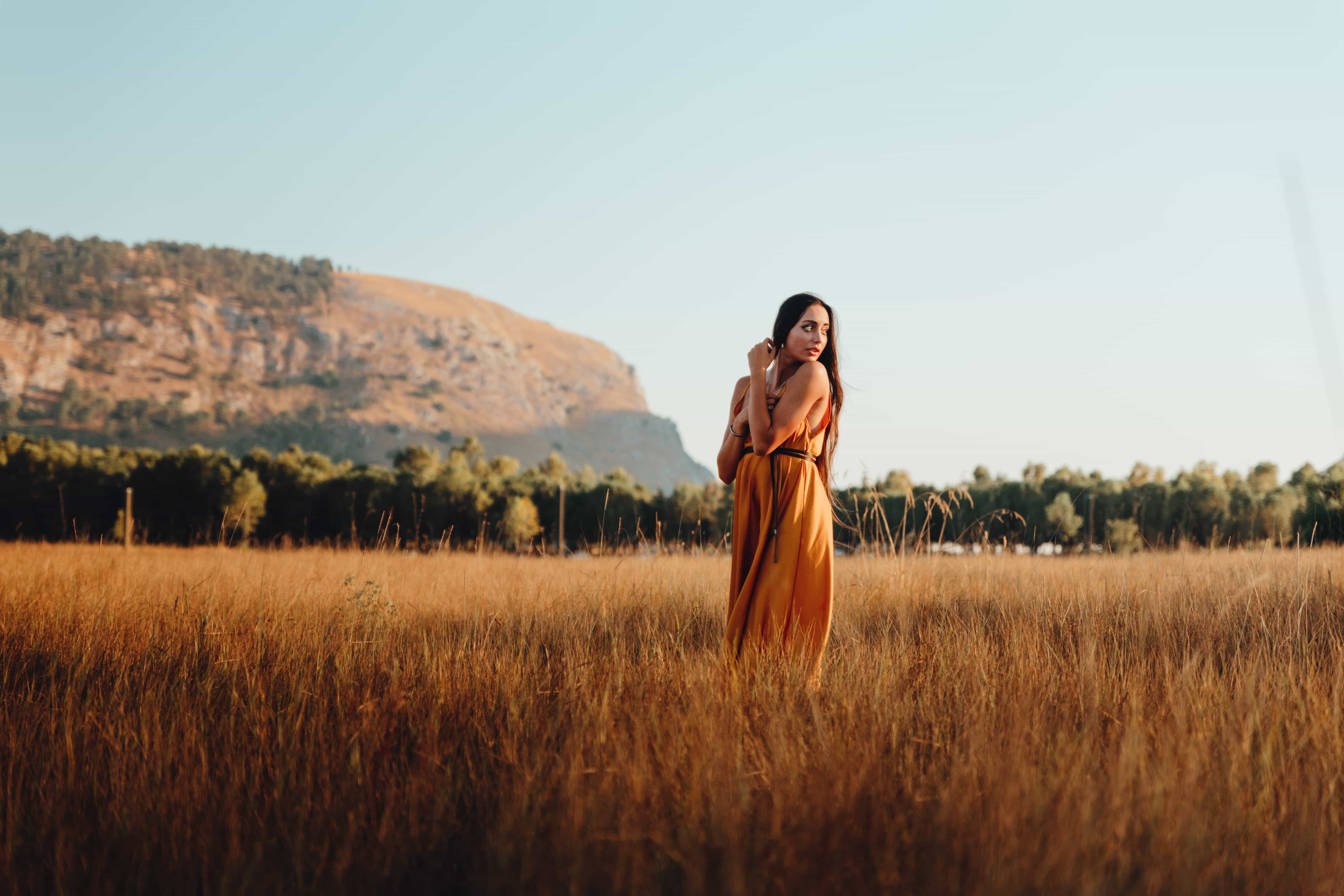 Woman in a yellow dress standing in a field with a mountain in the background, looking to the side
