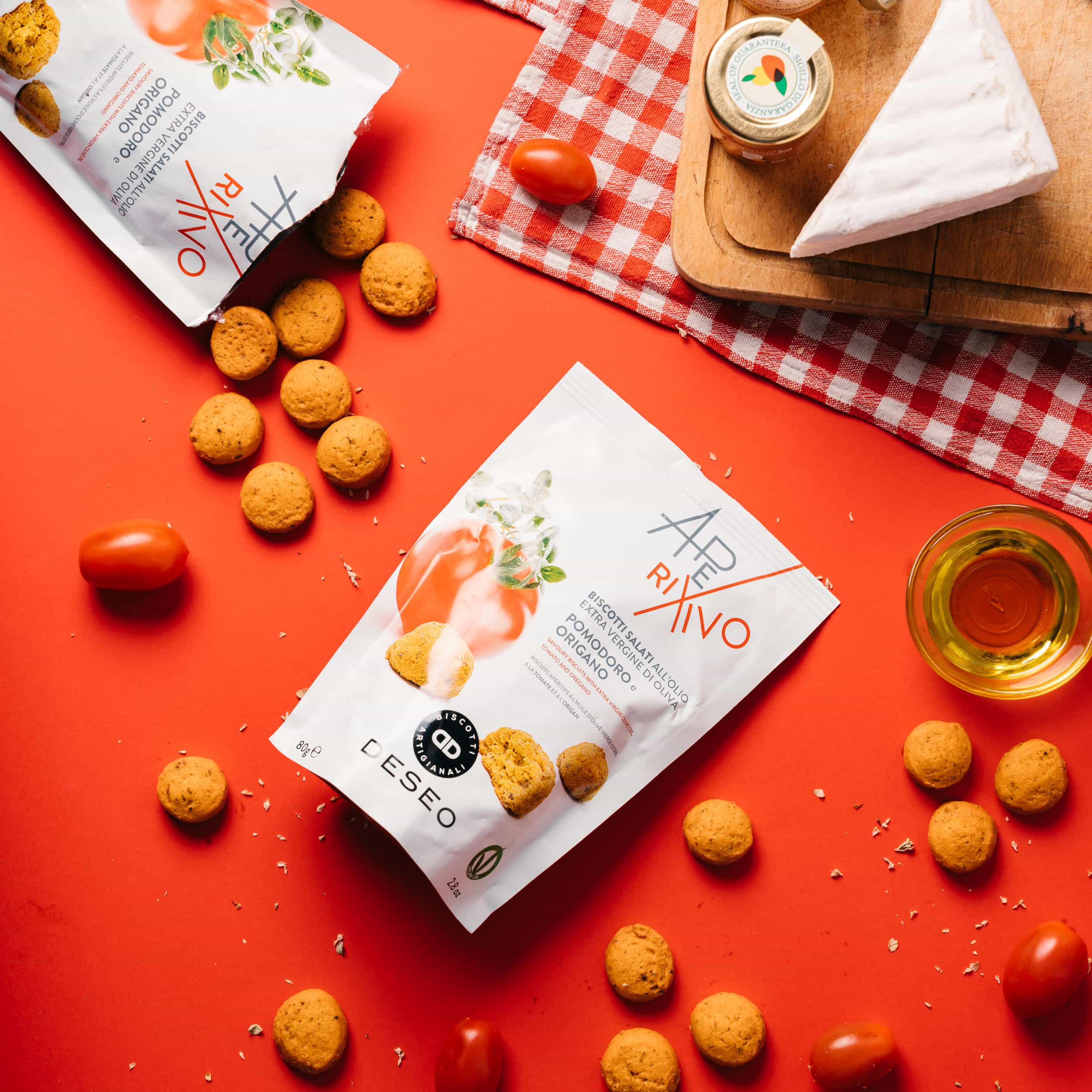 Two bags of Aperitivo snacks on a red surface with scattered snacks, cheese, tomato, and a glass of olive oil