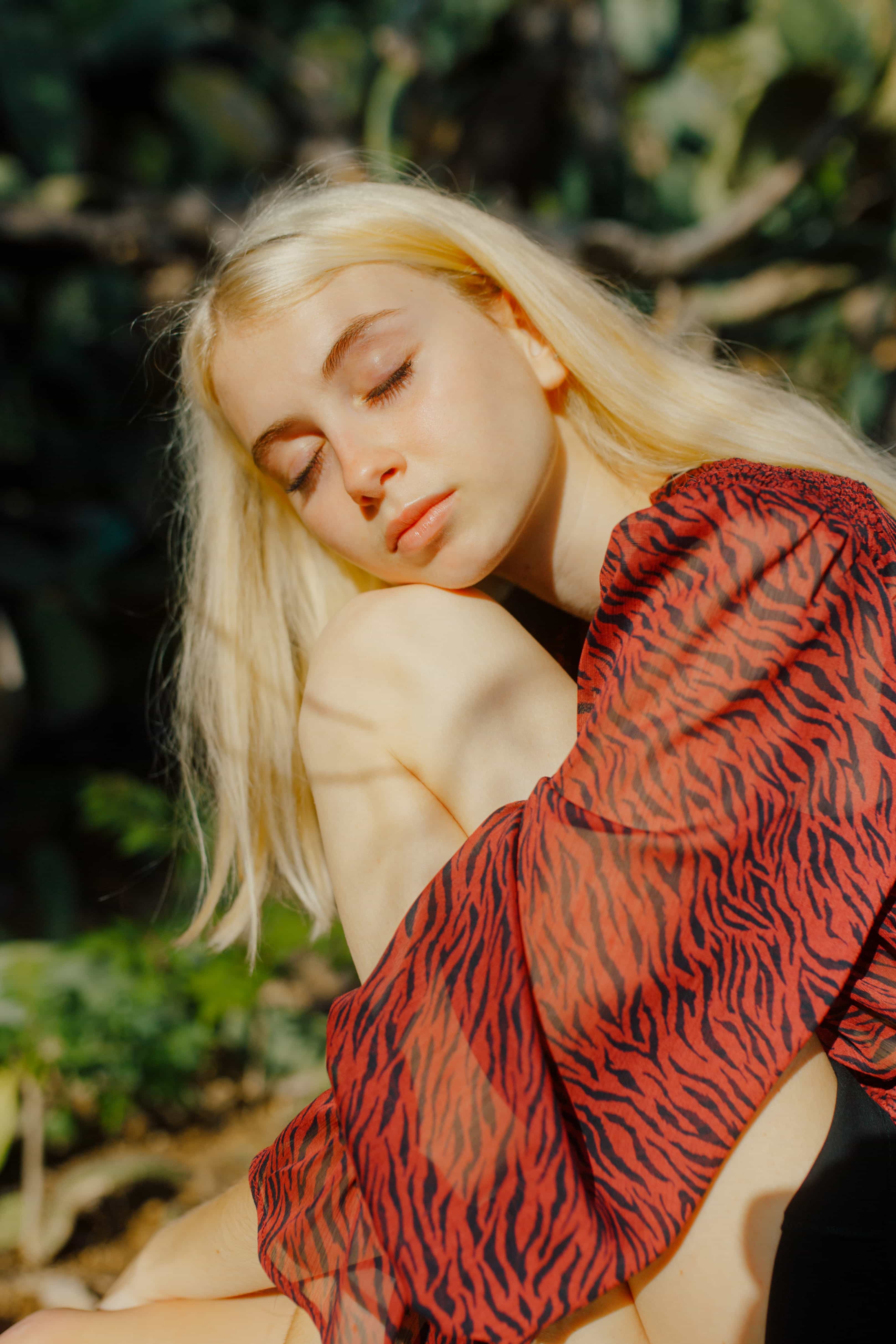 Portrait of a blonde woman with eyes closed, wearing a red and black patterned top, basking in sunlight