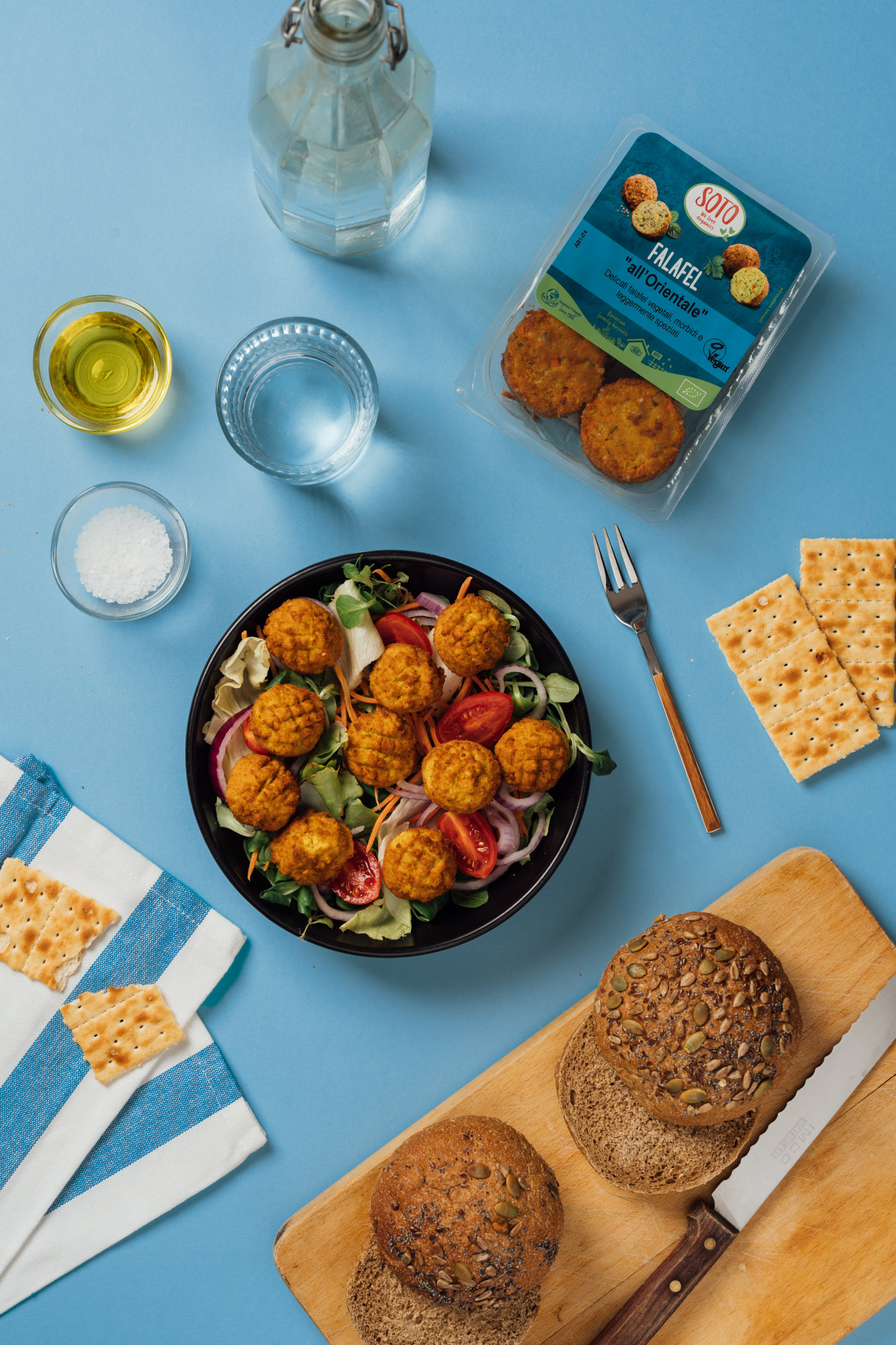 A top-down photo of a salad with falaffel, the product used a bottle of water and some crackers on a blue background