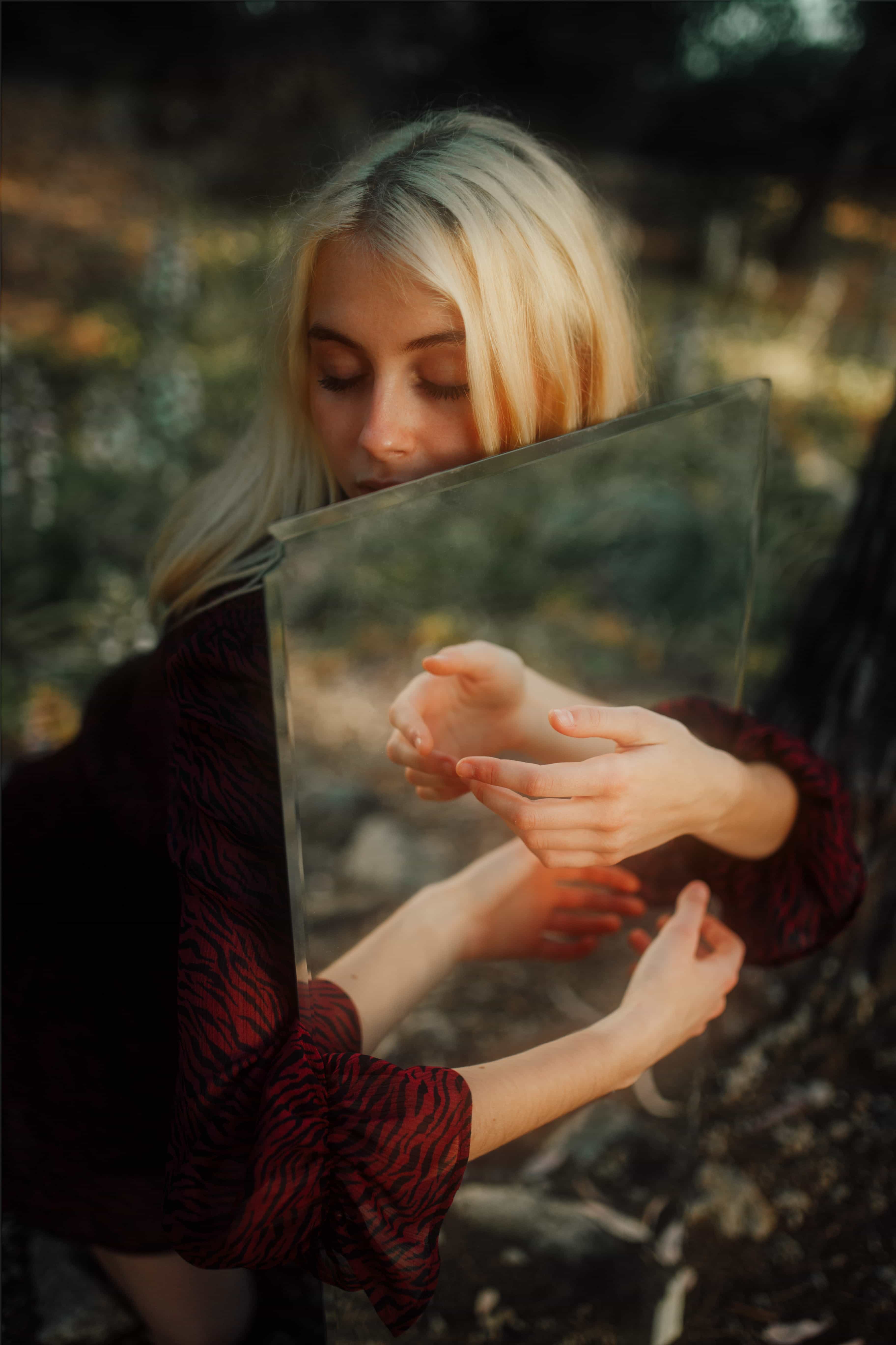 Blonde woman with eyes closed, holding a mirror reflecting her hands, set against a blurred outdoor background