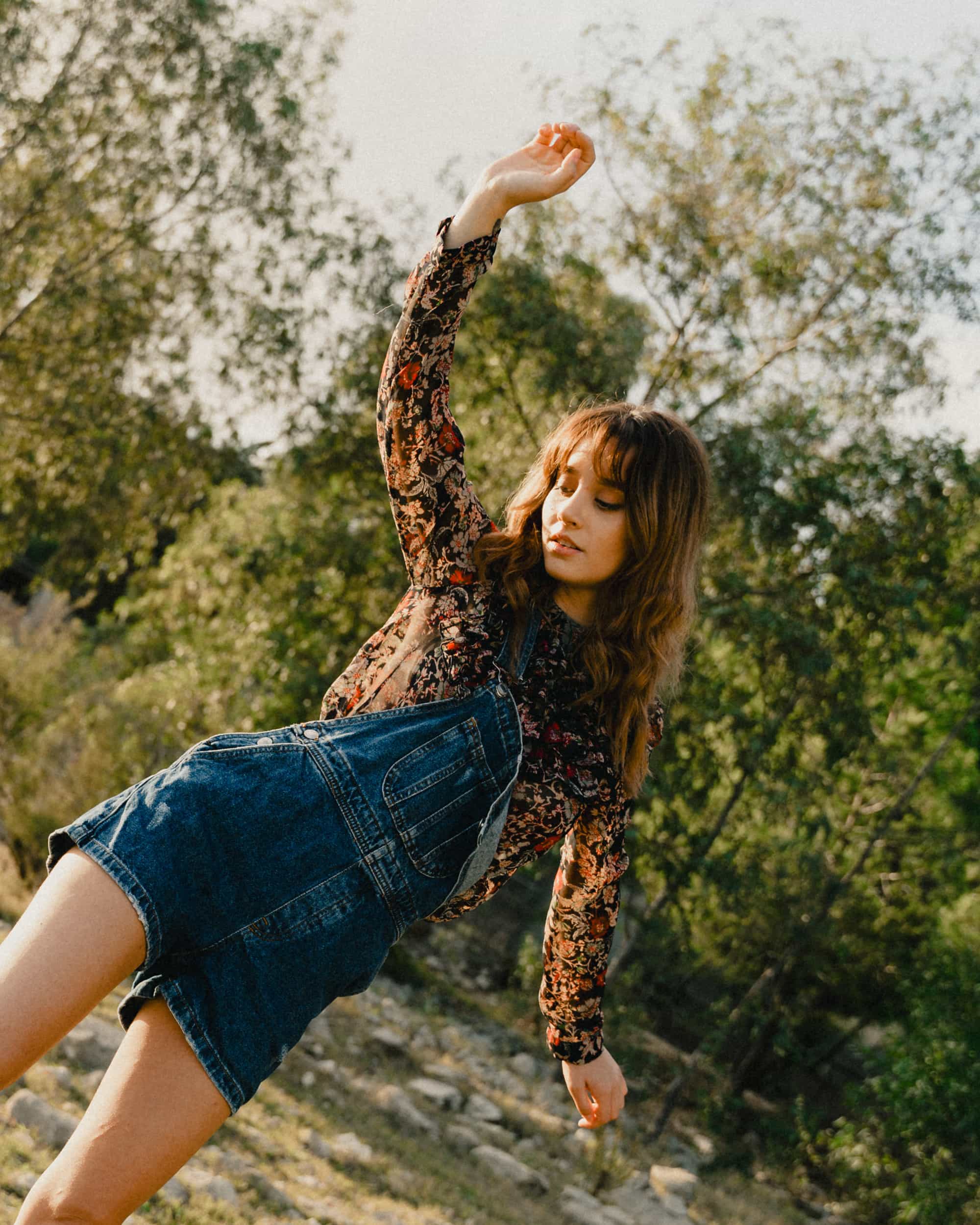 A woman wearing denim overalls and a patterned long-sleeve shirt raises her arm while standing outdoors with trees in the background.