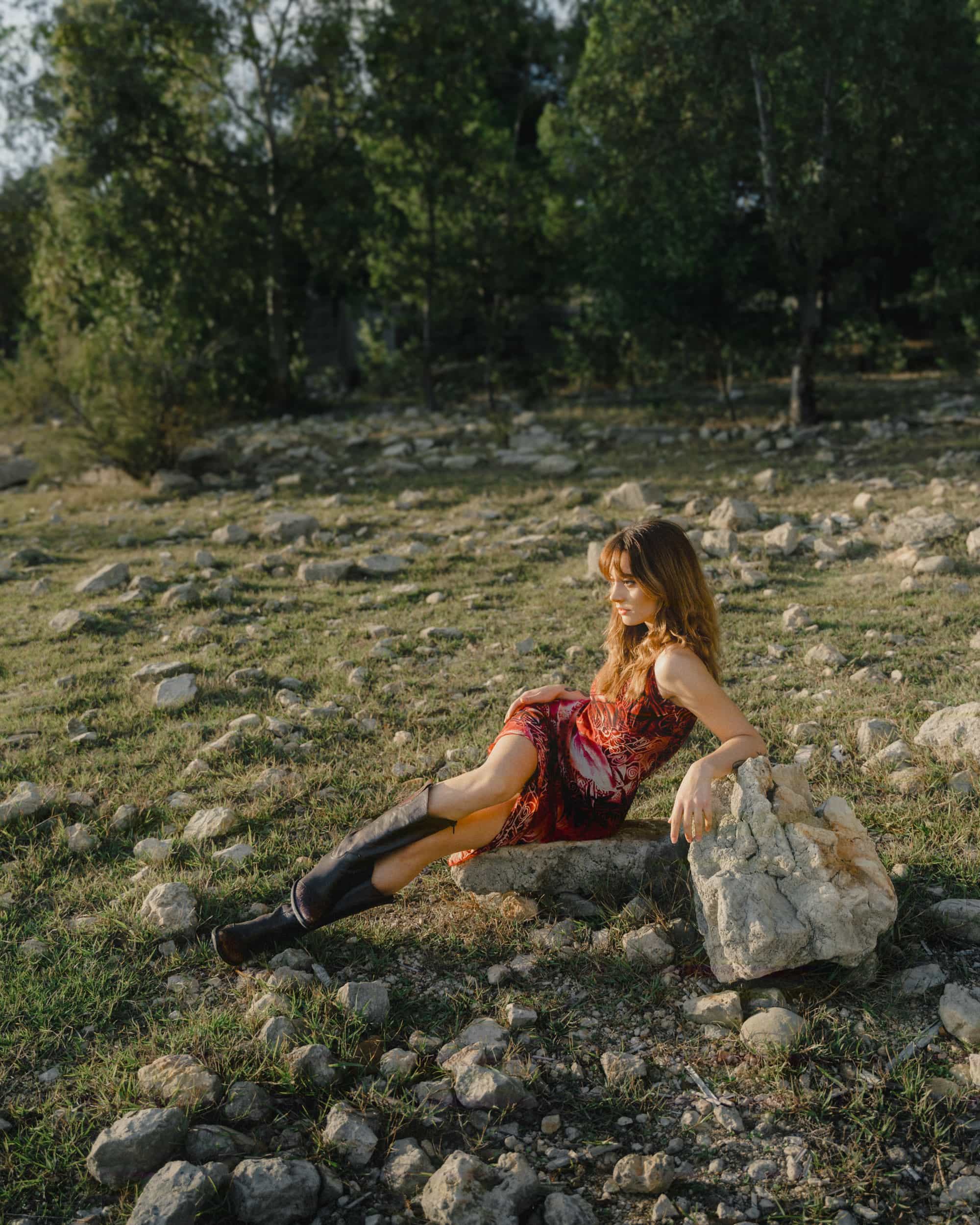 A person sits on a large rock in an outdoor setting, wearing a red dress and black boots. The background is filled with scattered rocks and trees.