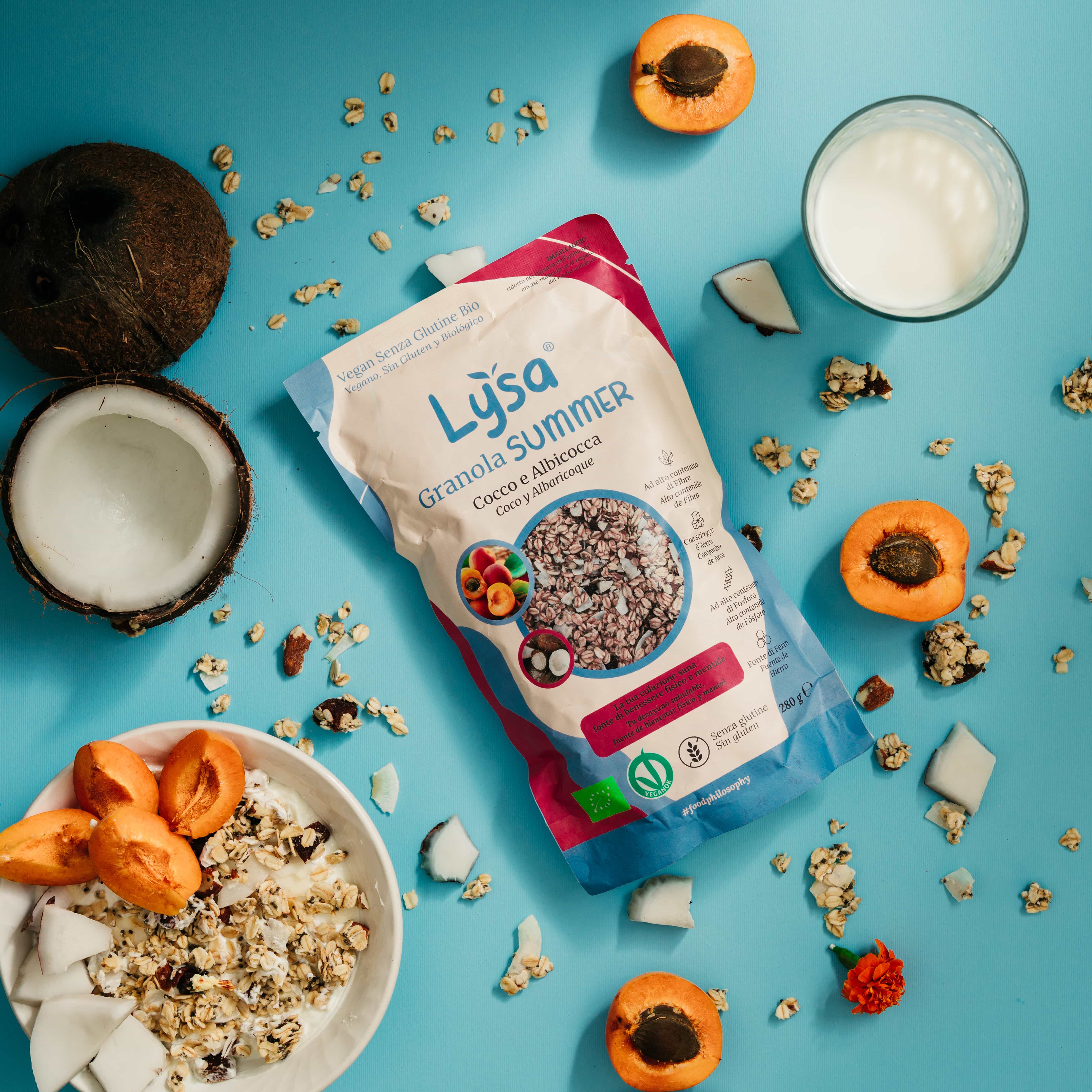 Package of Lysa Granola Summer with coconut and apricot flavor, surrounded by fresh apricots, coconut pieces, and a bowl of granola with milk. The scene is set on a light blue background with scattered granola and fruit.