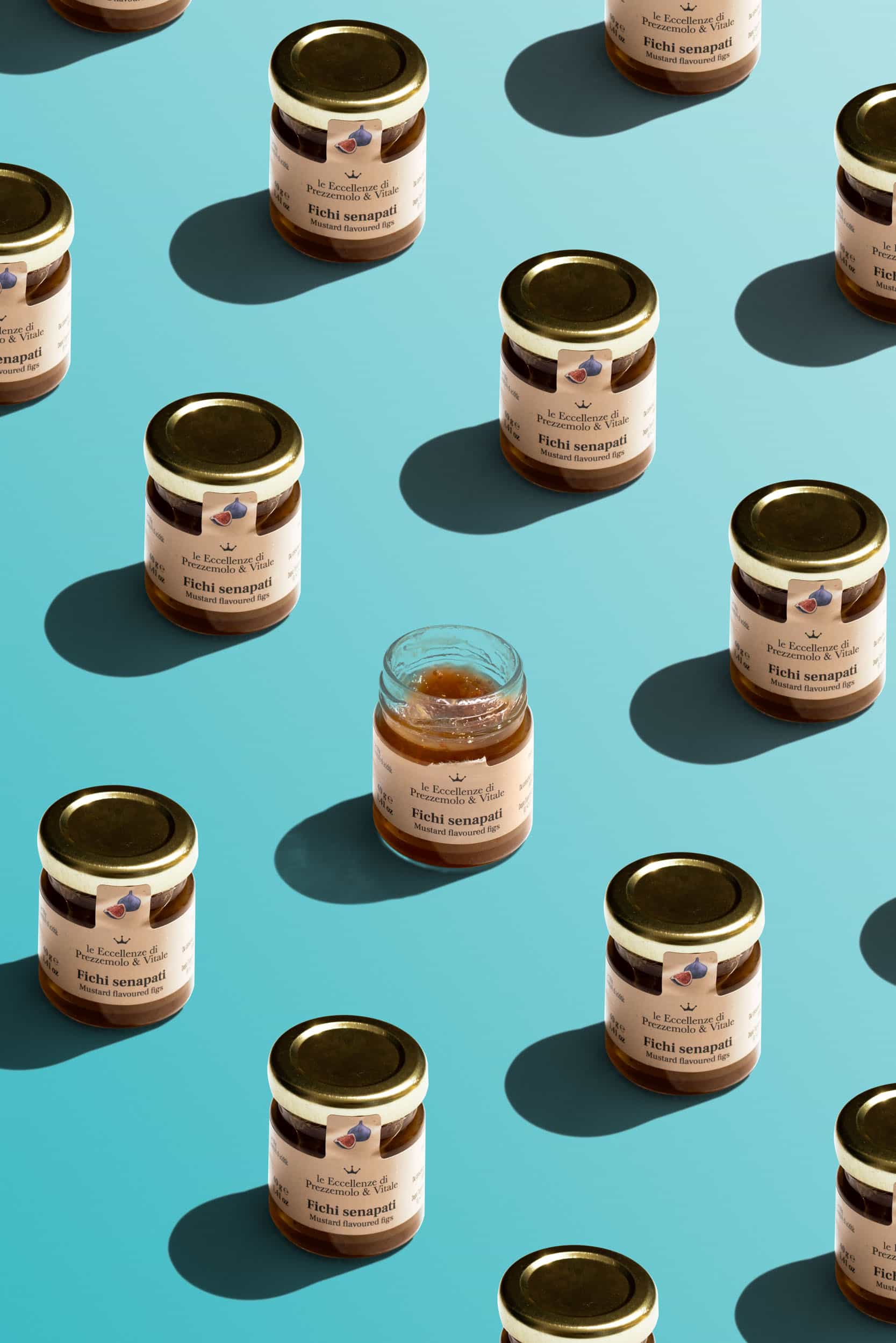 Multiple jars of 'Fichi Senapati' mustard-flavored fig jam arranged on a light blue background, with one jar open and partially used in the center.
