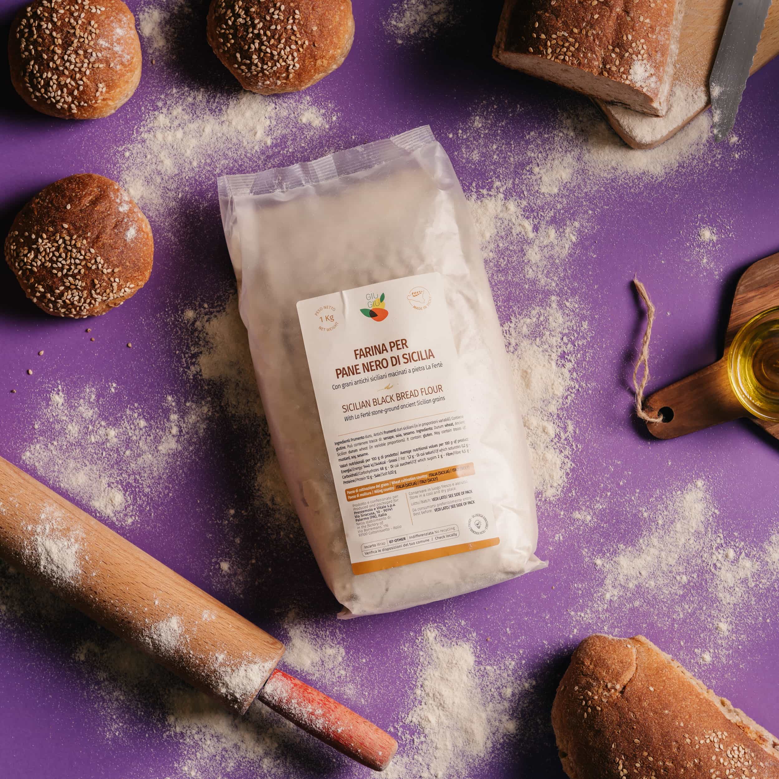 Package of Sicilian black bread flour on a purple surface with bread rolls, a loaf, rolling pin, and olive oil