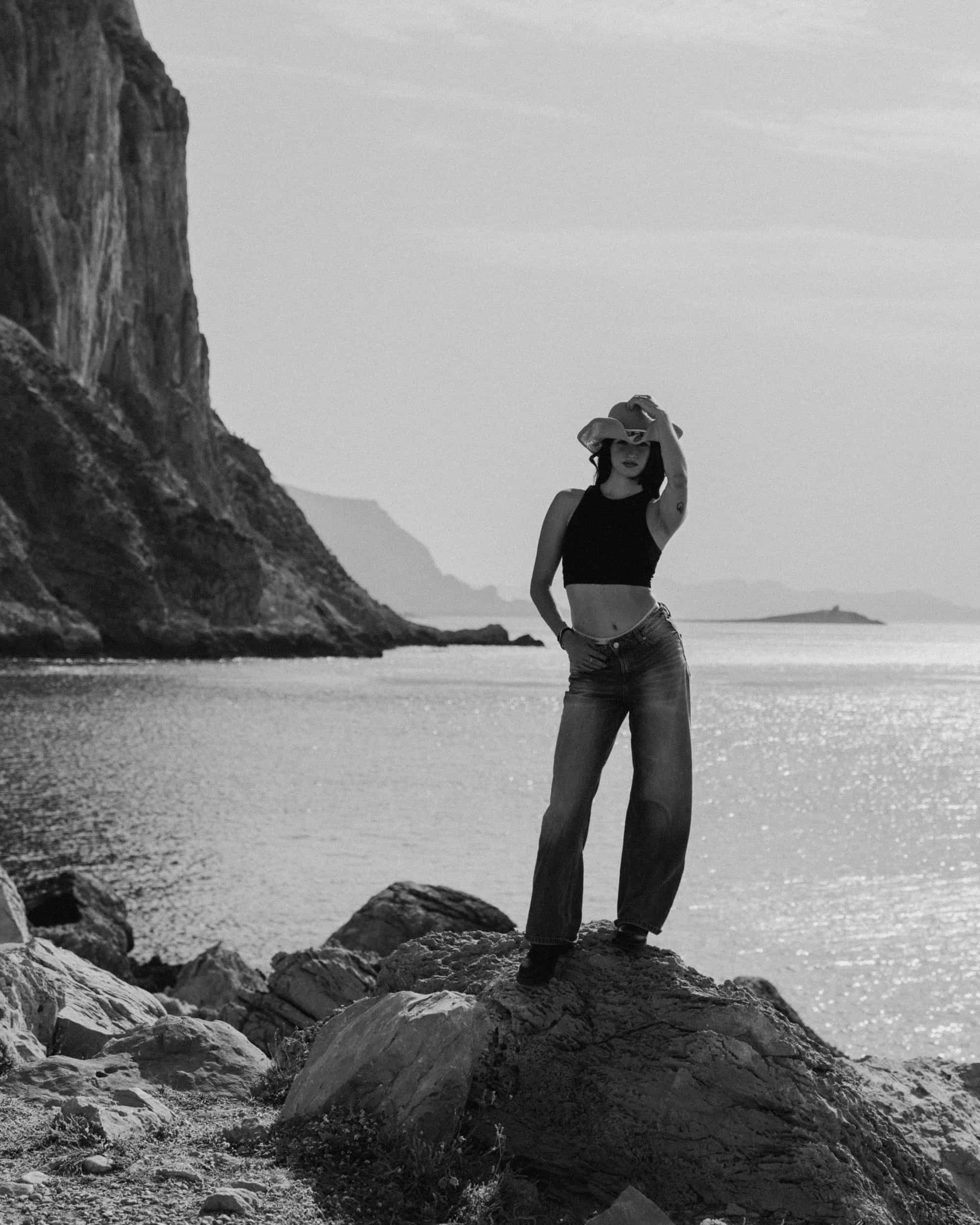 A woman in a cowboy hat standing on rocks near the ocean